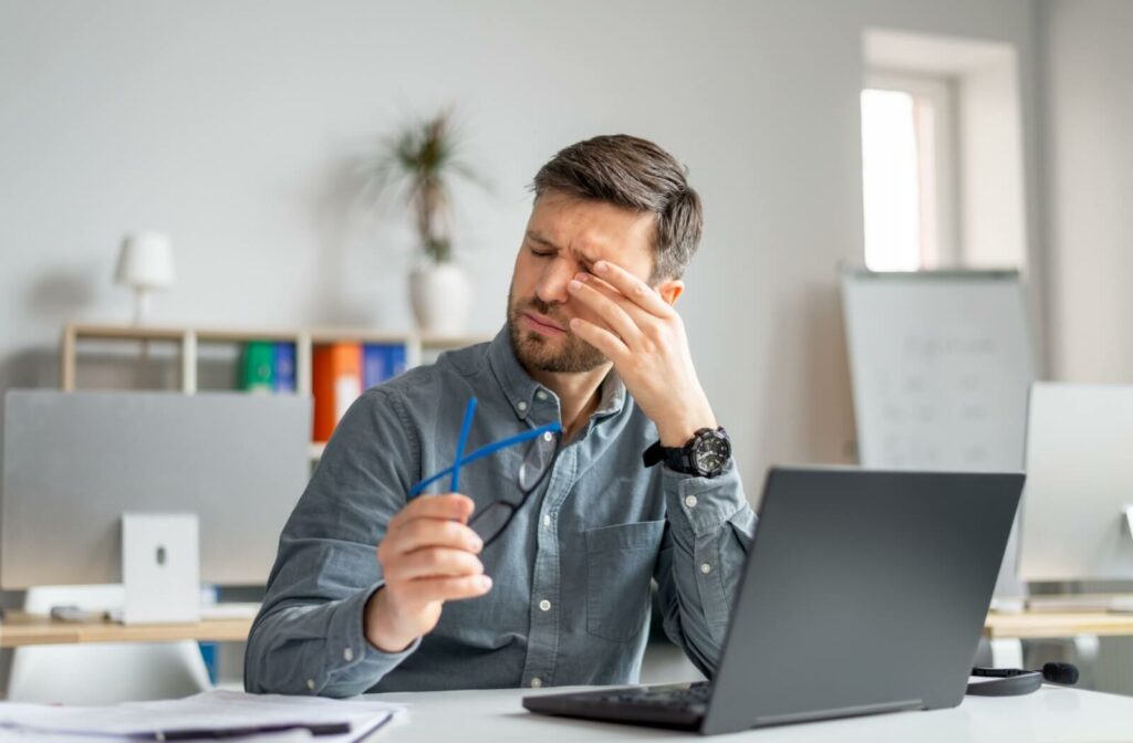 A person sitting at an office desk with a laptop, removing his glasses to rub his eyes in discomfort.