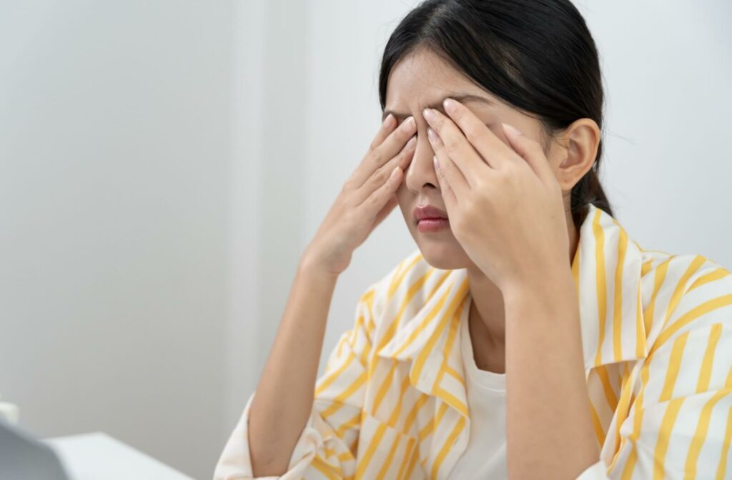 A person in a yellow striped shirt sitting at a desk and rubbing her closed eyes to relieve dryness and blurry vision.